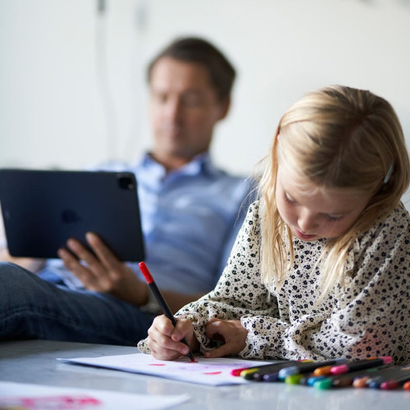 A child drawing in the foreground and a father using an iPad in the background.