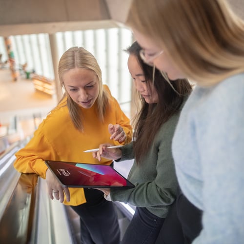 Tre damer står og ser ned på en ipad i en rulletrapp
